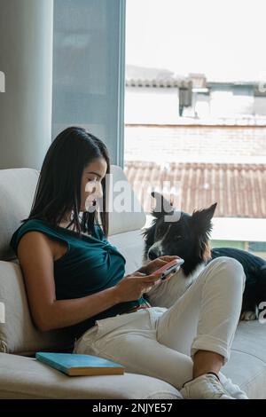 Young Hispanic female owner with long dark hair in casual clothes sitting on sofa next to adorable obedient Border Collie dog and messaging on smartph Stock Photo