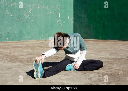 Focused female athlete in sportswear stretching legs while practicing Head to Knee pose on street Stock Photo