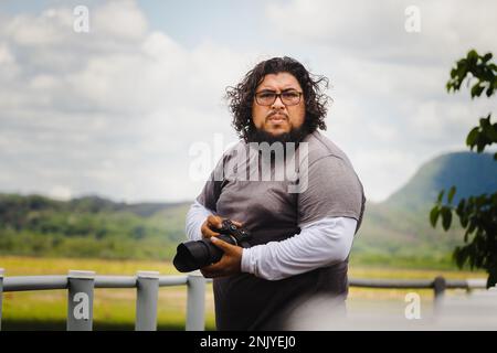 Serious bearded male in casual clothes and eyeglasses looking at camera while holding DSLR camera in countryside at Costa Rica Stock Photo