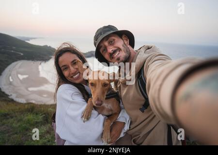 Smiling adult man and woman in casual clothes with backpacks standing on grassy hill with cute basenji dog and taking self portrait against sea at sun Stock Photo
