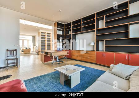 Interior design of contemporary living room with white sofa near red chair on blue rug with small coffee table and orange wooden workplace desk agains Stock Photo