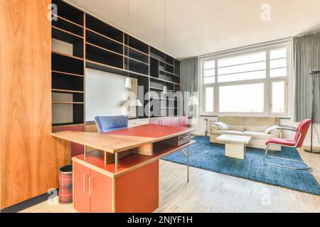 Interior design of contemporary living room with white sofa near red chair on blue rug with small coffee table and orange wooden workplace desk agains Stock Photo