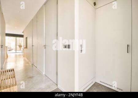 Minimalistic interior design of hall with white tall doors and walls with built in closet and light wooden bench in long narrow corridor with big wind Stock Photo