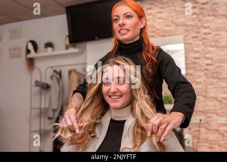 Cheerful female hairdresser smiling and checking hairstyle of happy young blond haired client in beauty salon Stock Photo