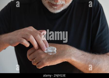 Crop unrecognizable bearded male in black t shirt applying cream on hand while standing against white wall Stock Photo