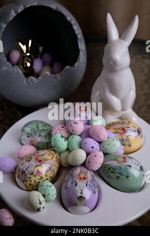 photo bright decorative large and small eggs lie on a special plate and next to a white rabbit on a round glass table Stock Photo