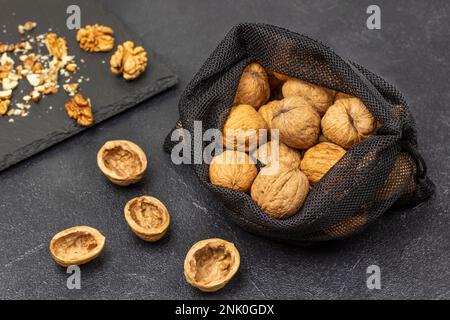 Walnuts in a reusable bag. Nut shells on the table, nut kernels on a black stone board. Black background. Stock Photo