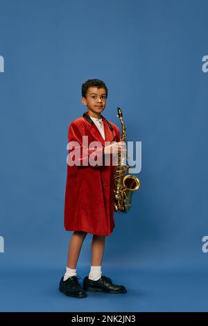 Portrait of cute little african boy wearing huge man's jacket and shoes like jazz man playing on saxophone over blue background. Fashion, art, music Stock Photo