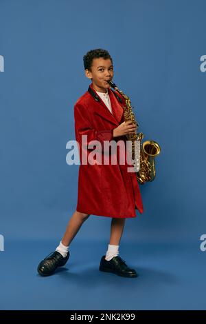 Portrait of cute little african boy wearing huge man's jacket and shoes like jazz man playing on saxophone over blue background. Fashion, art, music Stock Photo