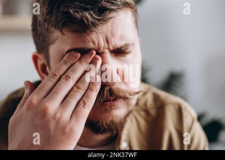 Selective focus of frustrated sad caucasian man suffering from strong eye pain. Eyesight problem. Upset young bearded male 30s touching eyes feeling d Stock Photo