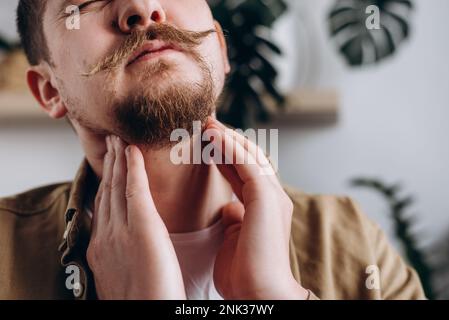 Close up of sad young bearded man holding hands over sore throat feeling discomfort. Painful neck and frowning, thyroid disorders, suffering sore thro Stock Photo