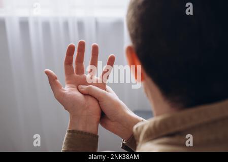 Close up side view of young caucasian male suffering from pain in palm. Ill man massaging painful hand from symptoms of peripheral neuropathy. Numbnes Stock Photo
