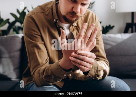 Selective focus of unhealthy tired young bearded man 30s with wrist pain tries to relieve suffering sitting on sofa in living room at home. Wrist male Stock Photo
