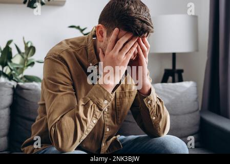 Close up of depressed young bearded man sitting on grey couch in living room, leaning on hands, having financial troubles during quarantine or sufferi Stock Photo