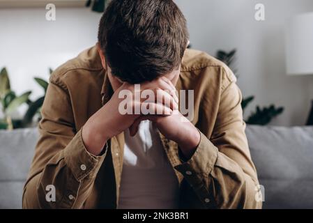 Depression and sadness concept. Close up of depressed young caucasian man sitting on sofa in living room, leaning on hands, having financial troubles Stock Photo