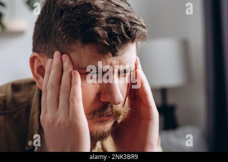 Closeup of upset young caucasian man suffering from headache sitting on couch at home, touching his temples. Migraine, headache, mental stress, panic Stock Photo