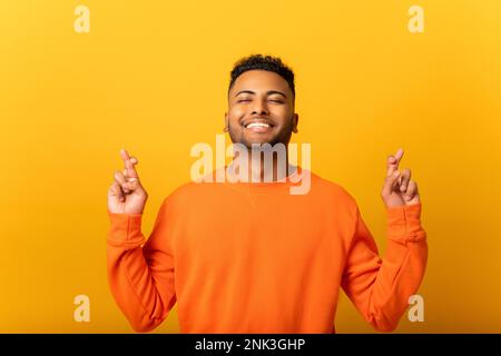 Wish you luck. Smiling young Indian man holding fingers crossed, looks up in hope, multiracial guy really wants to be lucky, isolated on yellow wall Stock Photo