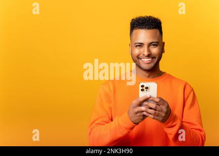 Photo of handsome cheerful smiling man holding mobile phone in the hand and smiling, isolated over yellow background, chatting using smartphone in front camera Stock Photo