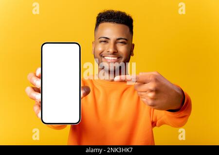 Surprised Indian man holding smartphone in hand and points finger at empty phone screen, astonishment guy presenting best deal, advertising new mobile app, isolated on yellow Stock Photo