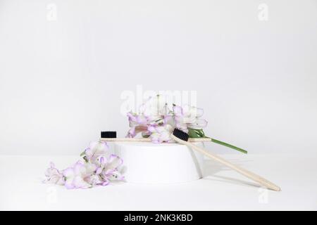 Creative representation of dentistry and oral care products. Bamboo toothbrushes on a podium pedestal with plant, flowers, on a white gray table backg Stock Photo