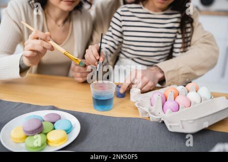 Cropped view of family holding paintbrushes near paints and Easter eggs in kitchen Stock Photo