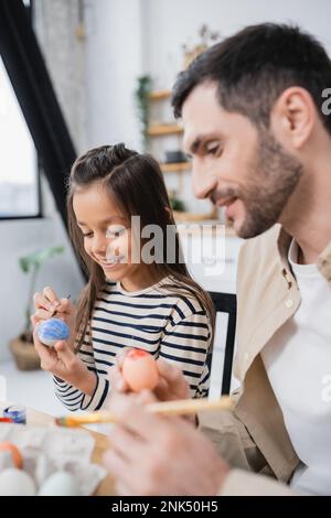Smiling girl painting Easter eggs near blurred father at home Stock Photo