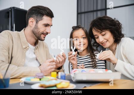 Smiling kid holding Easter rabbit figurine near parents and eggs at home Stock Photo