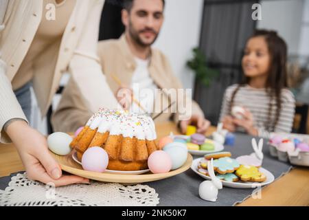 Woman putting plate with Easter cake on table near blurred family at home Stock Photo