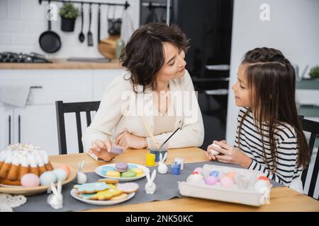 Smiling girl talking to mom near Easter eggs and sweet food on table Stock Photo