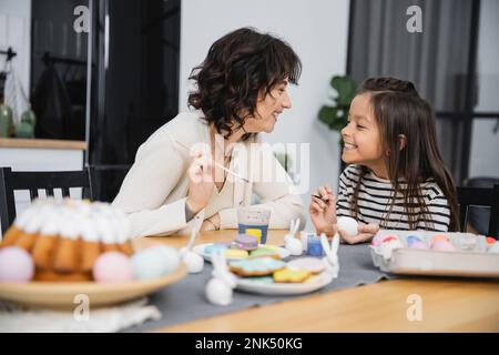 Positive mother and kid holding paintbrushes while coloring Easter eggs at home Stock Photo