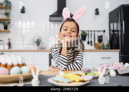 Cheerful girl in bunny ears headband sitting near Easter cake and eggs at home Stock Photo