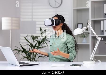 Successful latin american woman in office using vr glasses for video call and chatting online in virtual reality simulator, online conference with colleagues remote in metaverse. Stock Photo