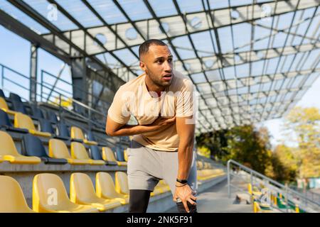 Young African American male sportsman, athlete, runner tired after training. He is standing in the stadium, bent over, holding his chest, breathing, resting. Stock Photo