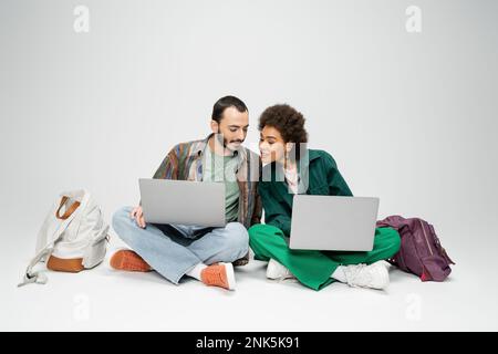 full length of interracial couple of students using laptops while sitting with crossed legs near backpacks on grey background,stock image Stock Photo