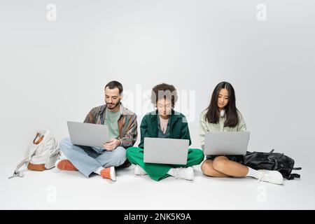 full length of multicultural students using laptops while sitting with crossed legs near backpacks on grey background,stock image Stock Photo