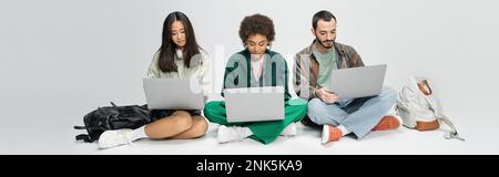 full length of stylish multiethnic students using laptops while sitting near backpacks on grey background, banner,stock image Stock Photo