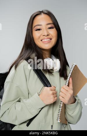 cheerful asian student with wireless headphones and copybook standing with backpack isolated on grey,stock image Stock Photo
