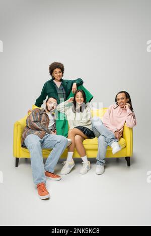 fashionable multiethnic friends posing on yellow couch and smiling at camera on grey background,stock image Stock Photo