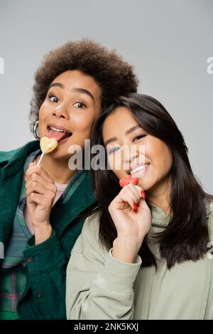 cheerful african american woman with lollipop sticking out tongue near asian friend isolated on grey,stock image Stock Photo