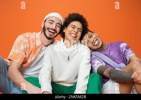 cheerful multiracial man looking at camera near friends laughing with closed eyes isolated on orange,stock image Stock Photo