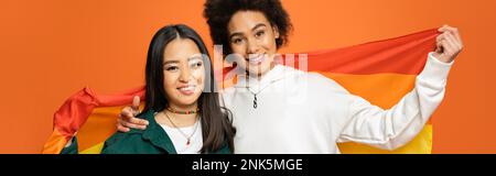 cheerful asian and african american women with lgbt flag looking at camera isolated on orange, banner,stock image Stock Photo