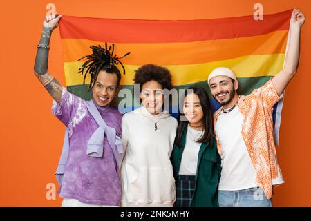 happy and trendy multiracial friends looking at camera while standing with lgbt flag isolated on orange,stock image Stock Photo
