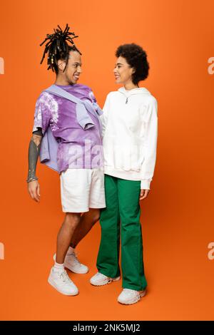 full length of cheerful african american woman and stylish man with dreadlocks looking at each other on orange,stock image Stock Photo