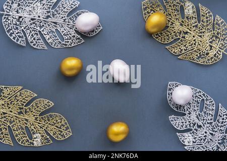 Gold and silver eggs, monstera leaves from wire on blue background. Stock Photo