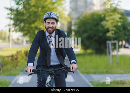 Smiling male employee in black suit commuting to work on bike lane in sunny morning. Front view of happy bearded worker looking at camera, while drivi Stock Photo