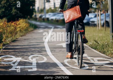 Unrecognizable male businessman in black suit riding on bicycle track in sleeping area of city. Crop view of office clerk with brown leather laptop ba Stock Photo