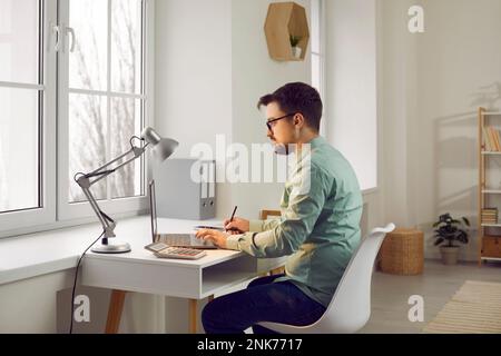 Attractive young man is sitting at his desk at home in the living room. Stock Photo