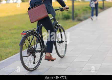 Back view of stylish man in suit cycling on bike on paved road outdoors. Crop view of unrecognizable businessman with brown leather briefcase riding o Stock Photo