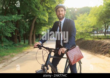 Male manager wearing suit, standing with bike on paved road, while getting to workplace. Front view of handsome man in helmet taking rest, while commu Stock Photo