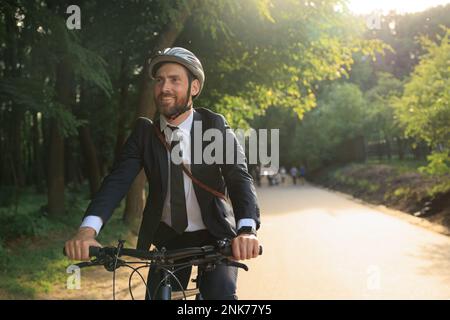Caucasian businessman in smart casual riding bicycle in park area to commute. Front view of smiling bearded man in blazer enjoying trip, while driving Stock Photo